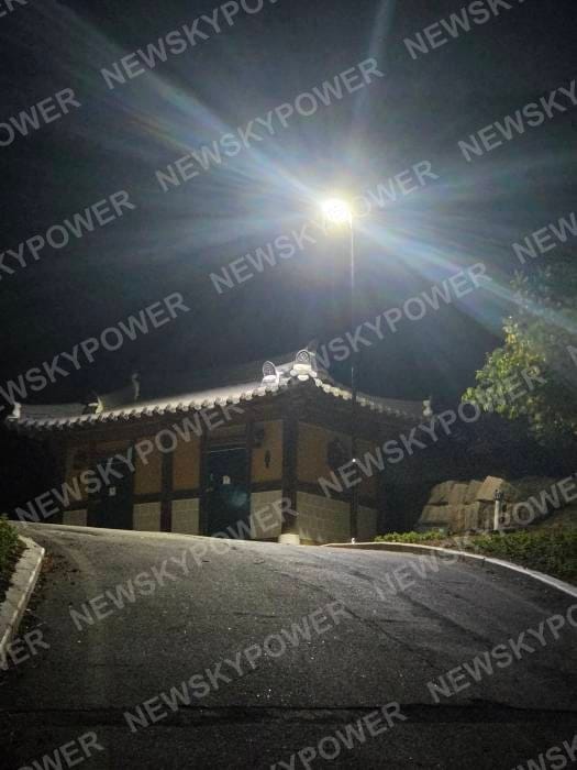 A tall light operating beside a building and sloping paved access area at night